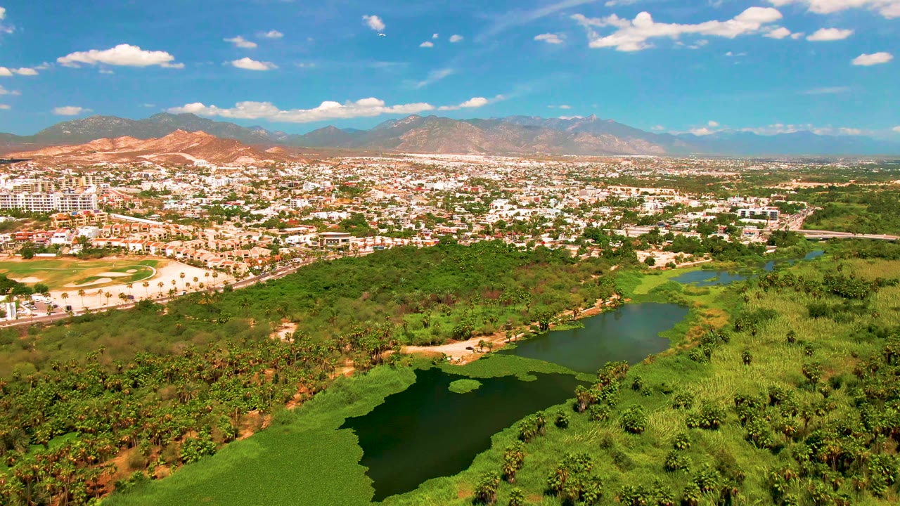 Opening aerial frame over San Jose Del Cabo with a green lake and dense vegetation in the foreground, city buildings beyond, reddish hills and distant mountains under scattered clouds, with no visible people.