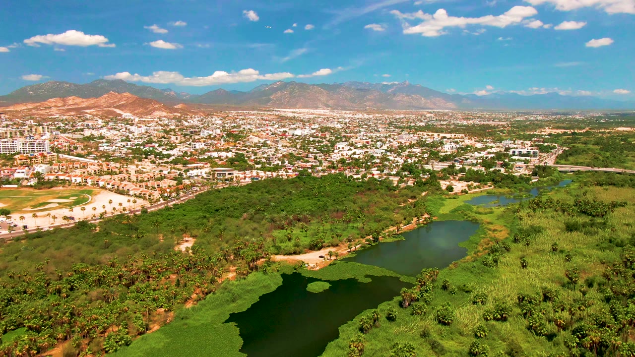 Aerial frame from San Jose Del Cabo showing the lake shifted lower in the foreground, green vegetation across the center, city blocks and roads in the distance, and mountains below a bright blue sky, with no visible people.