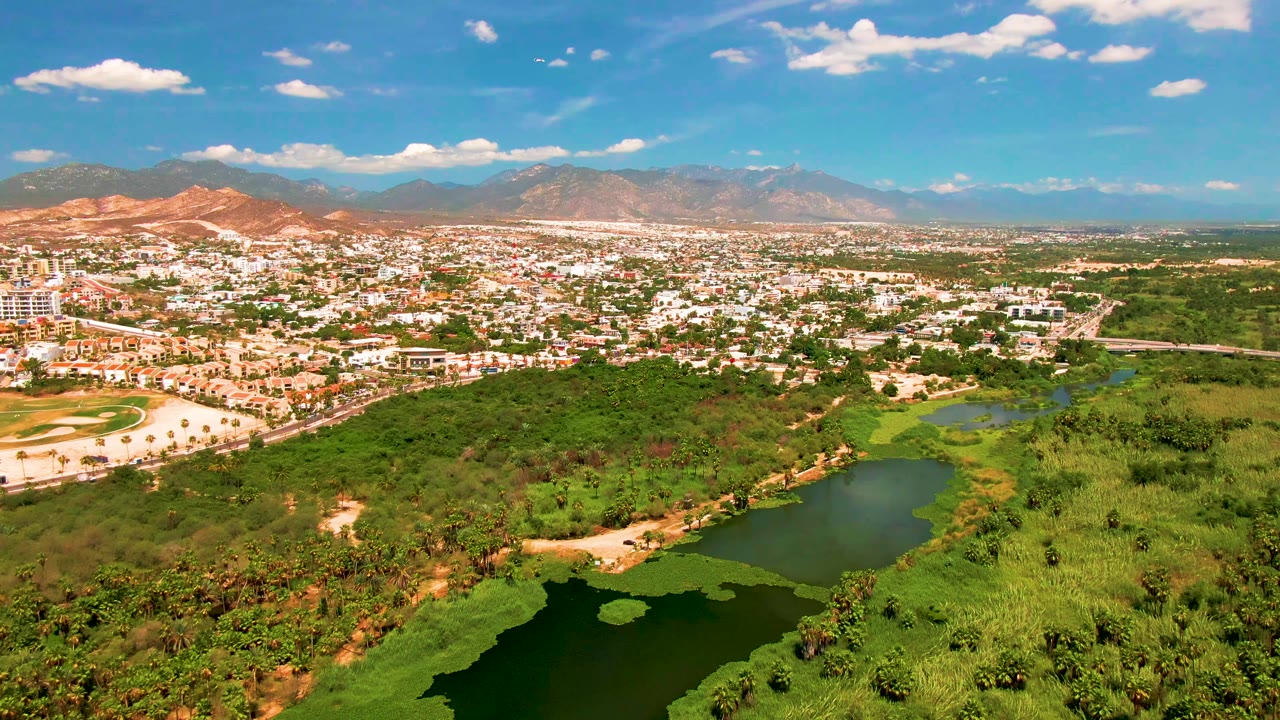 Aerial view of San Jose Del Cabo with dark green water at the bottom edge, bright vegetation, a low-rise city district, a road corridor, reddish hills, and distant mountains, with no visible people.