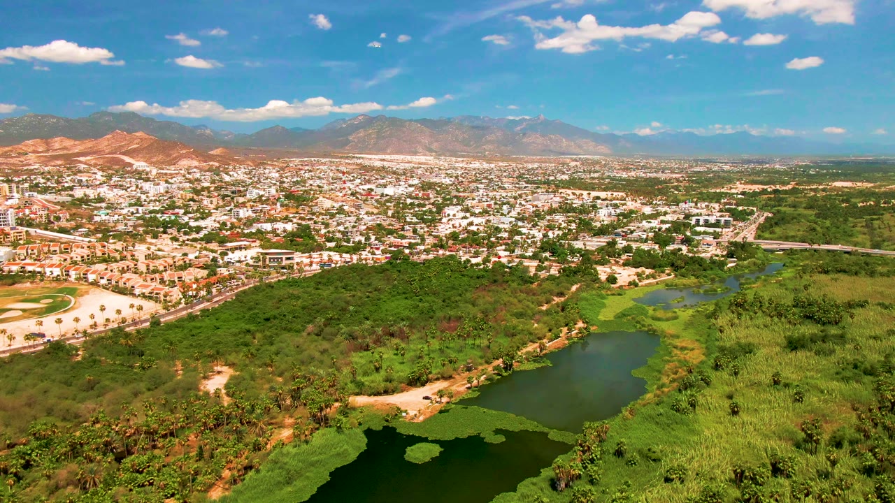 Mid-clip aerial frame over San Jose Del Cabo with the city edge and roads more centered, green wetland vegetation and lake below, reddish hills to the left, and hazy mountains in the background, with no visible people.