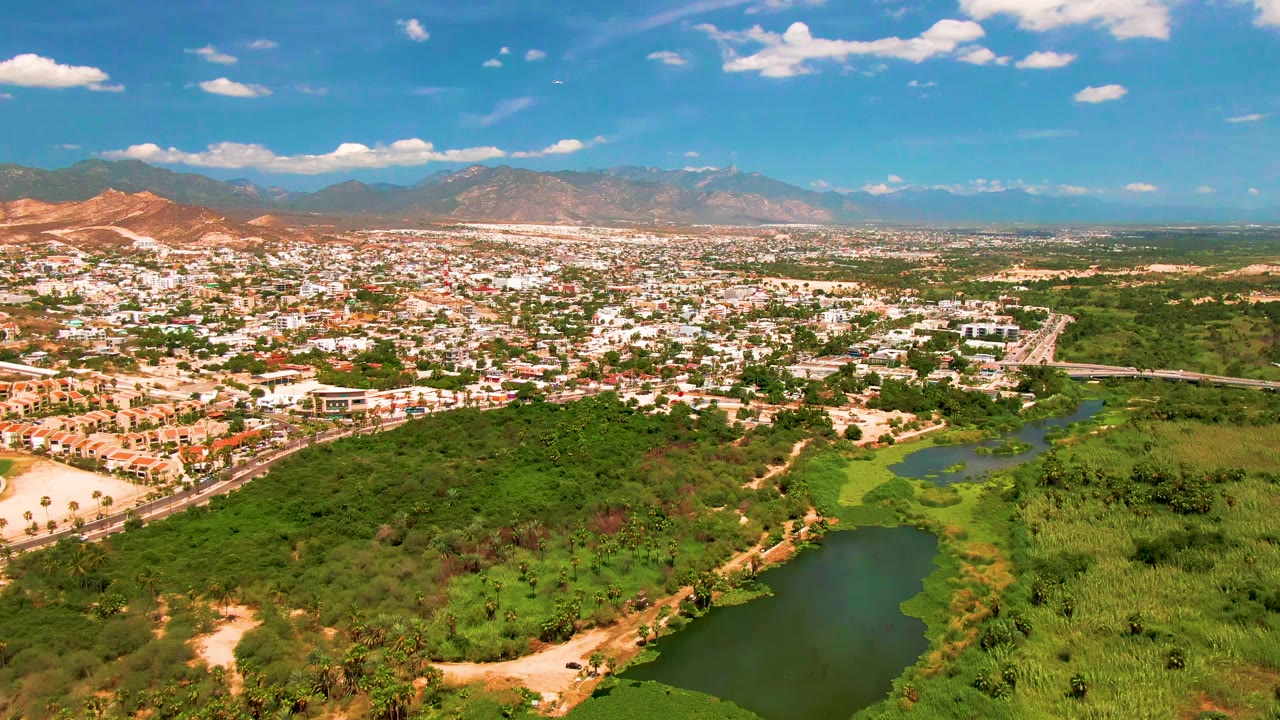 Wide aerial frame over San Jose Del Cabo, Mexico, showing a green river channel in the foreground, city buildings across the middle distance, mountains on the horizon, and no visible people.