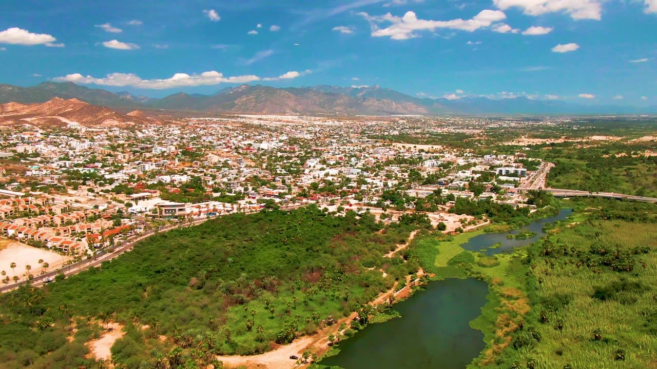 Drone view of urban buildings, river greenery, roads, hills, and distant mountains.