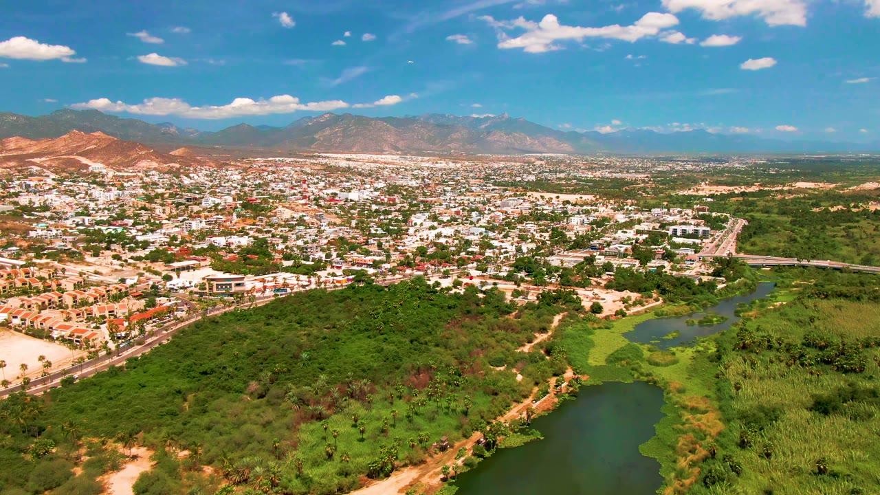 San Jose Del Cabo appears from above with dense green vegetation around the waterway, light-colored neighborhoods beyond it, a road crossing the scene, mountains in the background, and no visible people.