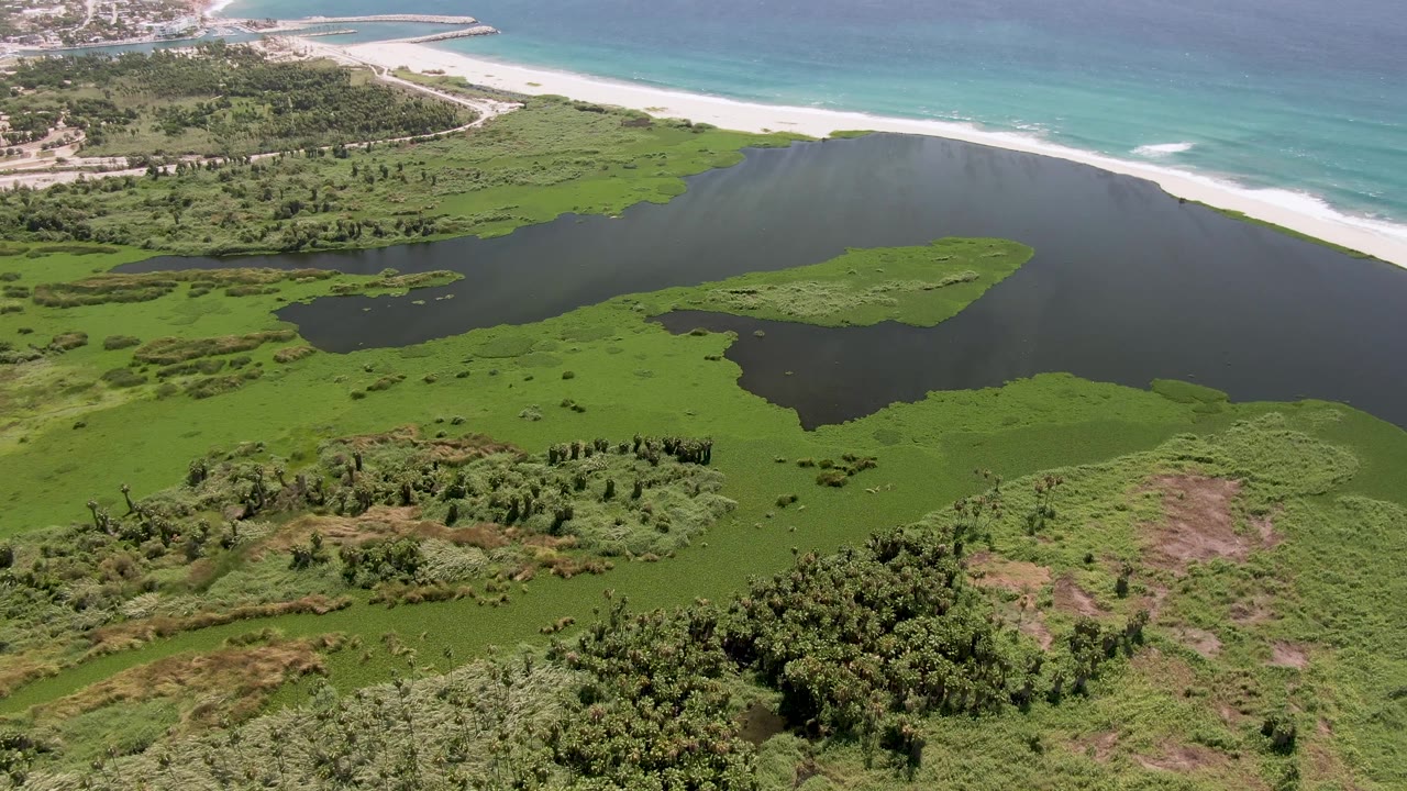 Opening aerial frame over San Jose Del Cabo wetlands with dark lagoon water, green marsh islands, a white sand beach, turquoise ocean, and no visible people.