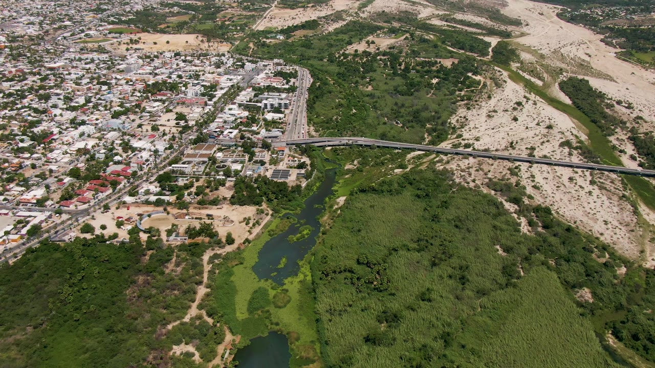 High aerial frame over San Jose del Cabo showing a bridge and road crossing beside a green river corridor, dense buildings, pale hills, and no visible people.