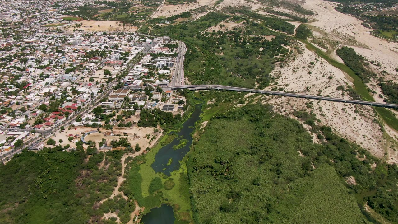 Drone perspective over San Jose del Cabo showing the roadway and bridge spanning the green river corridor, with residential streets to one side, sandy hills to the other, and no visible people.