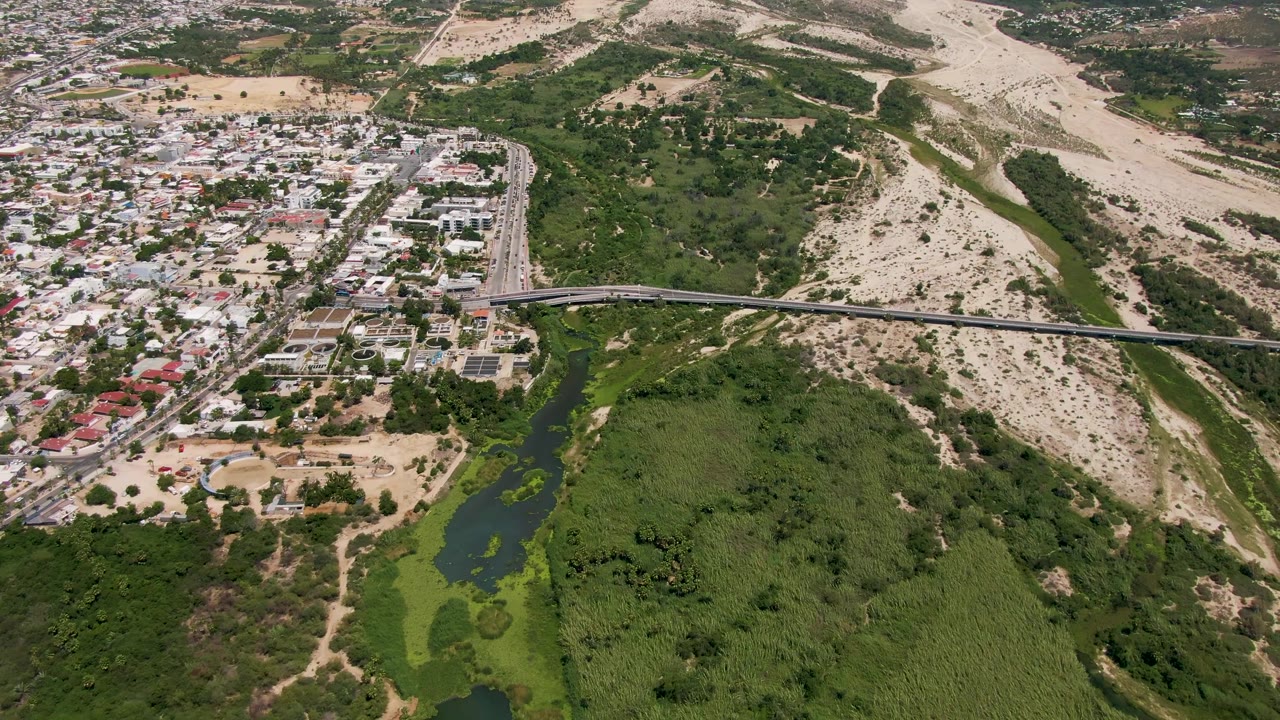 High aerial frame over San Jose Del Cabo showing a bridge across a green river corridor, nearby homes, pale hillside terrain, and no visible people.