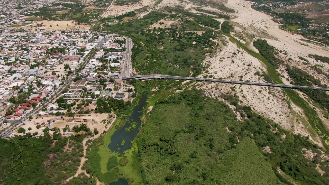 Aerial view in San Jose Del Cabo with the bridge centered over the river channel, town buildings to the left, dry hills beyond, and no visible people.