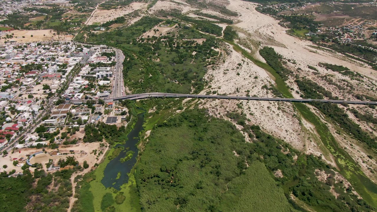 Aerial frame from San Jose Del Cabo showing a bridge over a green river corridor, roadside buildings, pale dry hills, and no visible people.