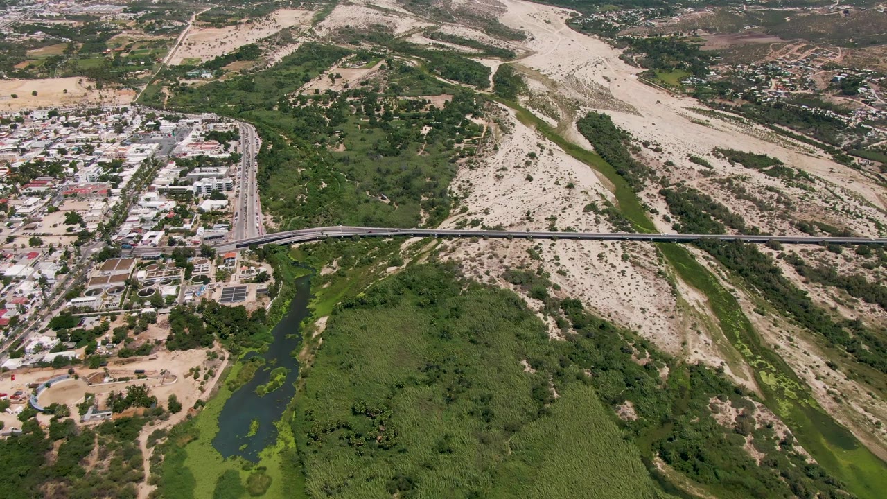 At 0:02, the frame places the bridge across the center with San Jose Del Cabo buildings to one side, dry hillside behind, and no visible people.
