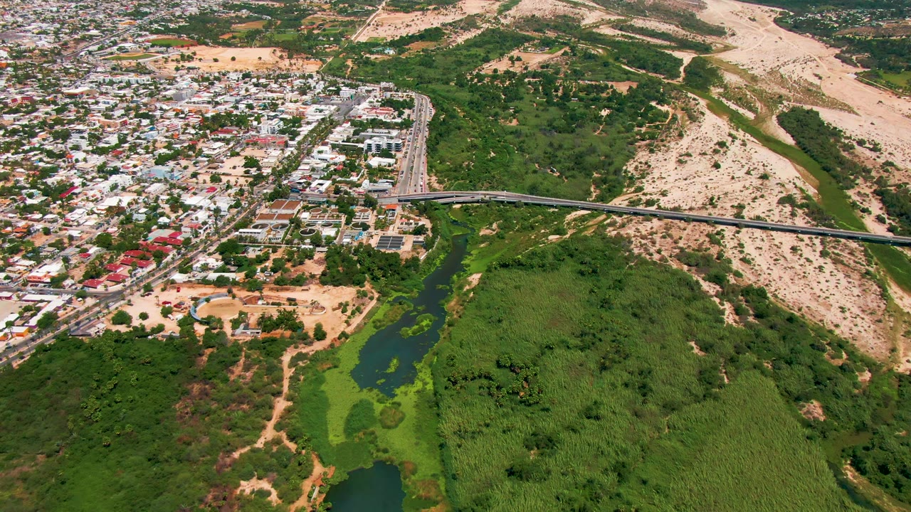 Opening aerial frame over San Jose del Cabo, Baja California Sur, with an overpass crossing green vegetation and water beside dense low-rise buildings and dry hills, with no visible people.