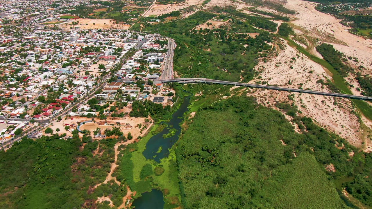 Drone view of vehicles on a bridge with town blocks, water, vegetation, and pale hills around it.