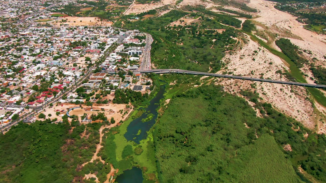 Aerial frame at 0:02 over San Jose del Cabo, with the bridge spanning the center, small vehicles visible on the roadway, dark water in the lower left, dense green vegetation, and dry hills in the background, with no visible people.