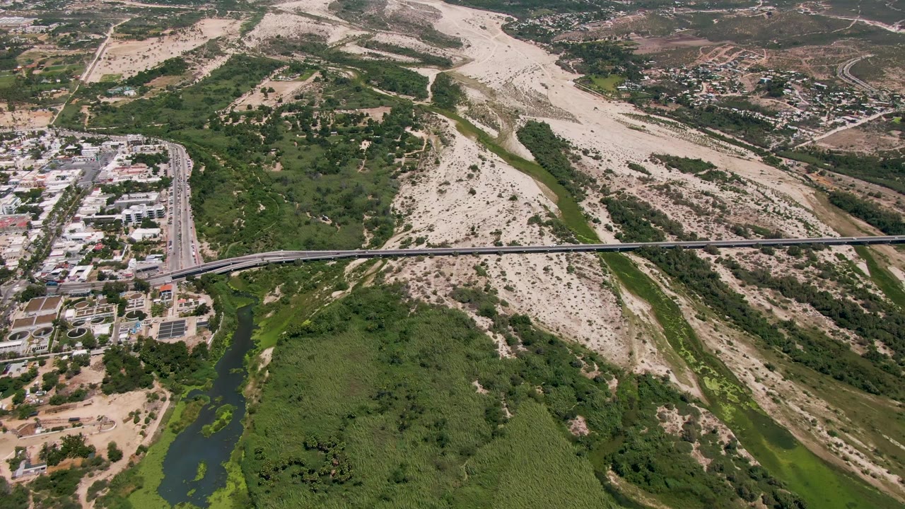 Opening aerial frame over San Jose Del Cabo, Baja California Sur, with a bridge crossing a green river corridor, pale dry hills beyond, buildings at left, and no visible people.