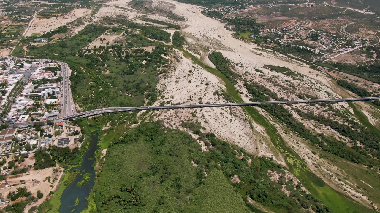 Aerial frame in San Jose Del Cabo showing the bridge stretched across the river channel, green vegetation below, light-colored dry terrain behind it, roadside buildings, and no visible people.