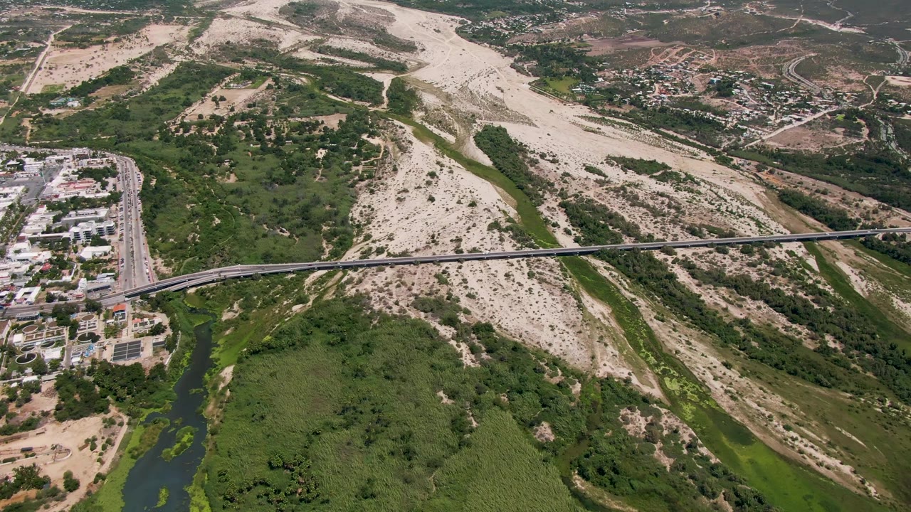 Mid-clip aerial frame over San Jose Del Cabo with the bridge crossing the middle of the image, the river and green banks below, buildings near the road, dry hills in the background, and no visible people.