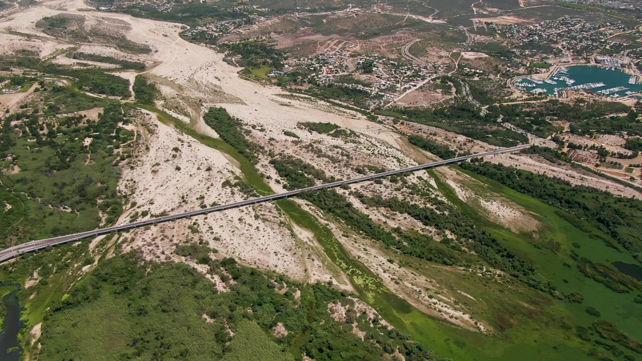 Aerial frame over San Jose del Cabo showing a pale dry riverbed, a green river channel, a diagonal bridge, nearby buildings, hills, and no visible people.