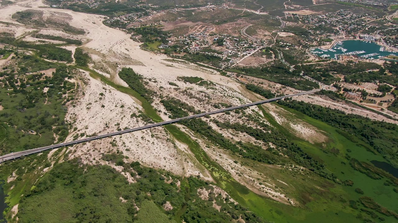 Drone view of a bridge crossing a pale riverbed with green water and vegetation below.