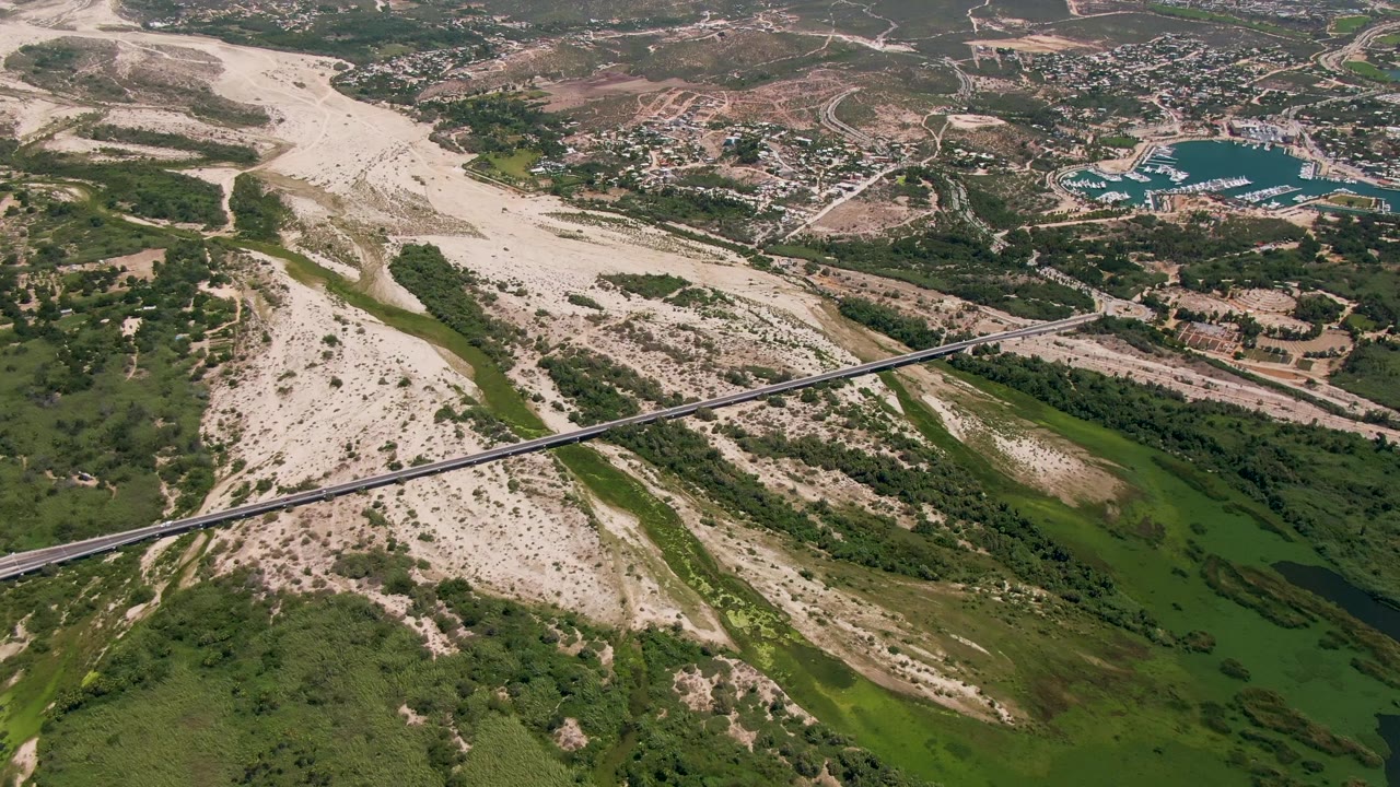 San Jose del Cabo aerial frame showing the river corridor running through a pale wash, the bridge angled across the scene, a small blue reservoir, scattered development, and no visible people.