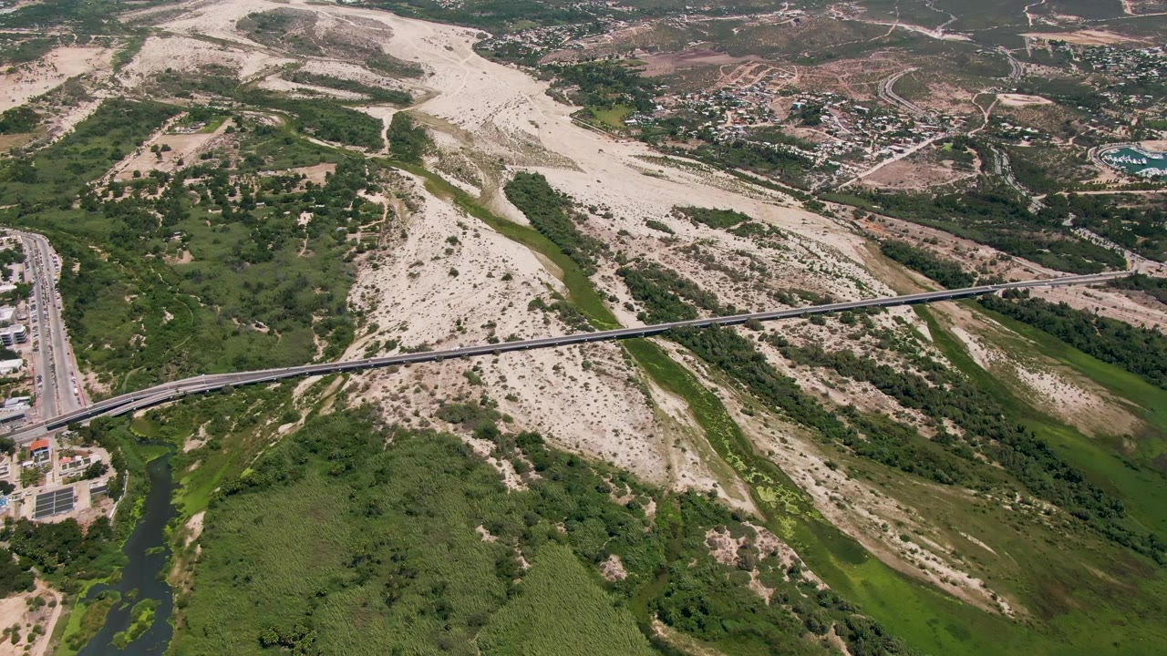 Aerial frame over San Jose Del Cabo, Baja California Sur showing a pale dry riverbed, a diagonal bridge, green wash vegetation, roads, distant buildings, and no visible people.