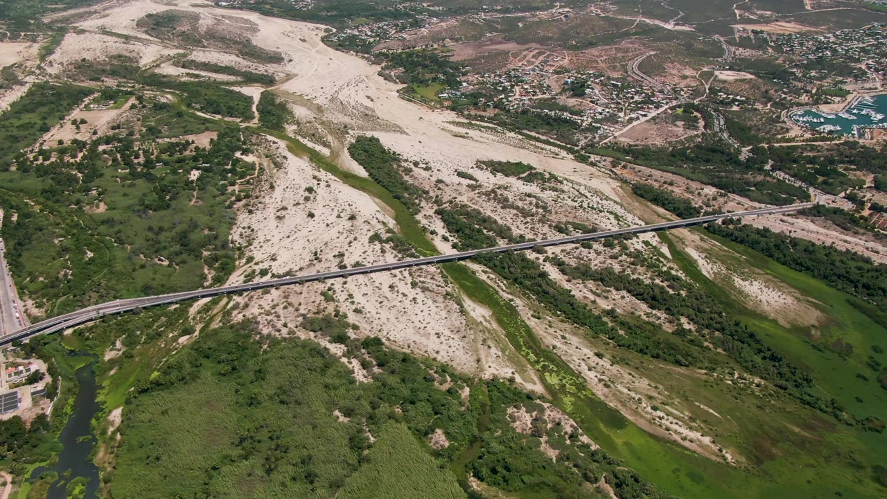 San Jose Del Cabo aerial frame showing the bridge spanning a dry riverbed, darker green vegetation and water in the lower wash, hillside roads, distant buildings, and no visible people.