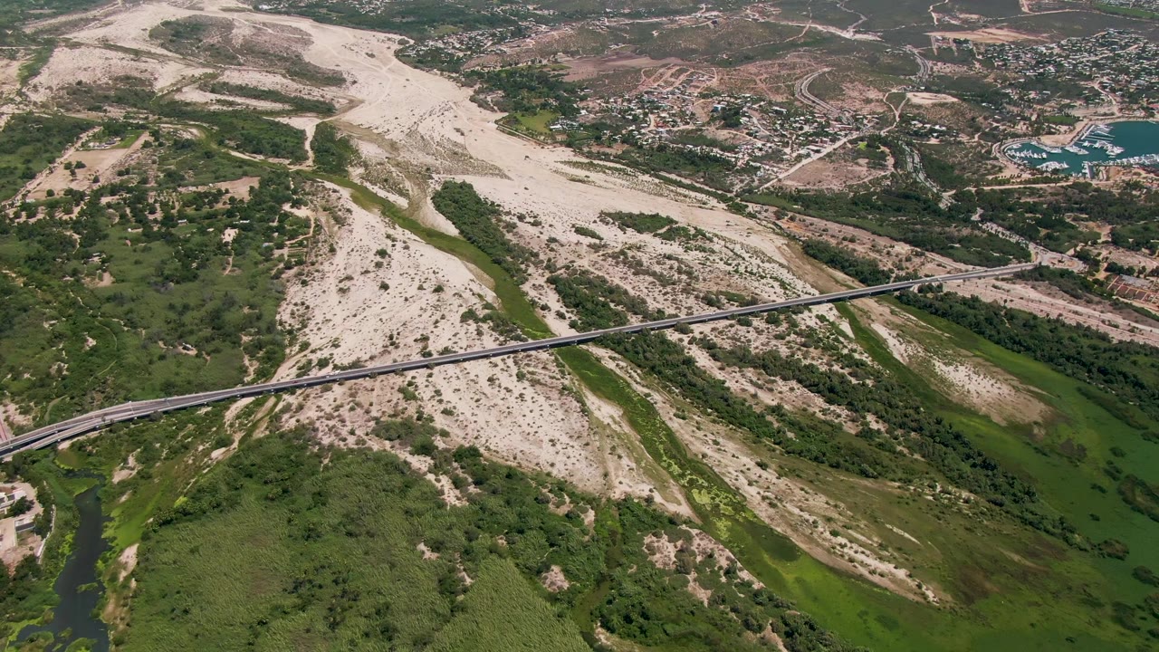 Aerial frame of a bridge crossing a dry riverbed with greenery and buildings beyond.