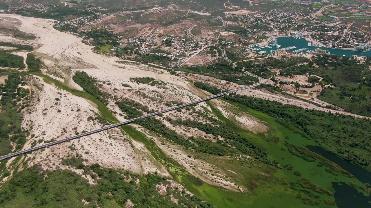 High aerial view over San Jose Del Cabo showing a green river corridor, pale sandy wash, diagonal bridge, low buildings, and blue coastal water in the distance, with no visible people.