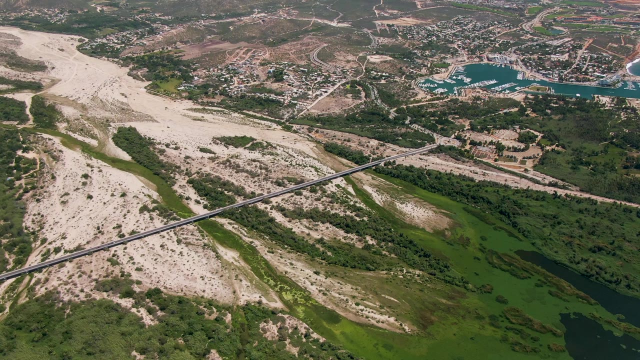 San Jose Del Cabo aerial frame with the diagonal bridge crossing the sandy riverbed, dense green vegetation below, and town blocks and turquoise water set farther back, with no visible people.