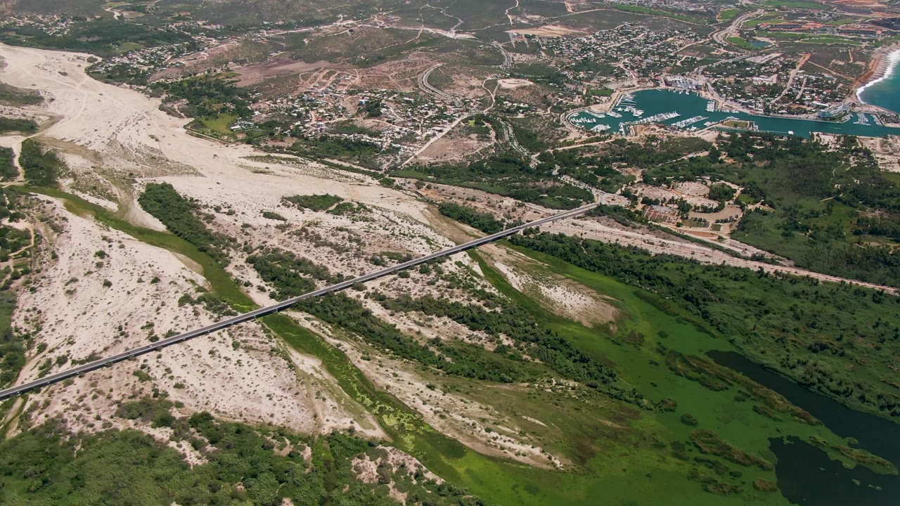Wide San Jose Del Cabo aerial view showing green wetlands and dark river water in the lower frame, a pale sand corridor and bridge in the middle, and beach and blue water at upper right, with no visible people.