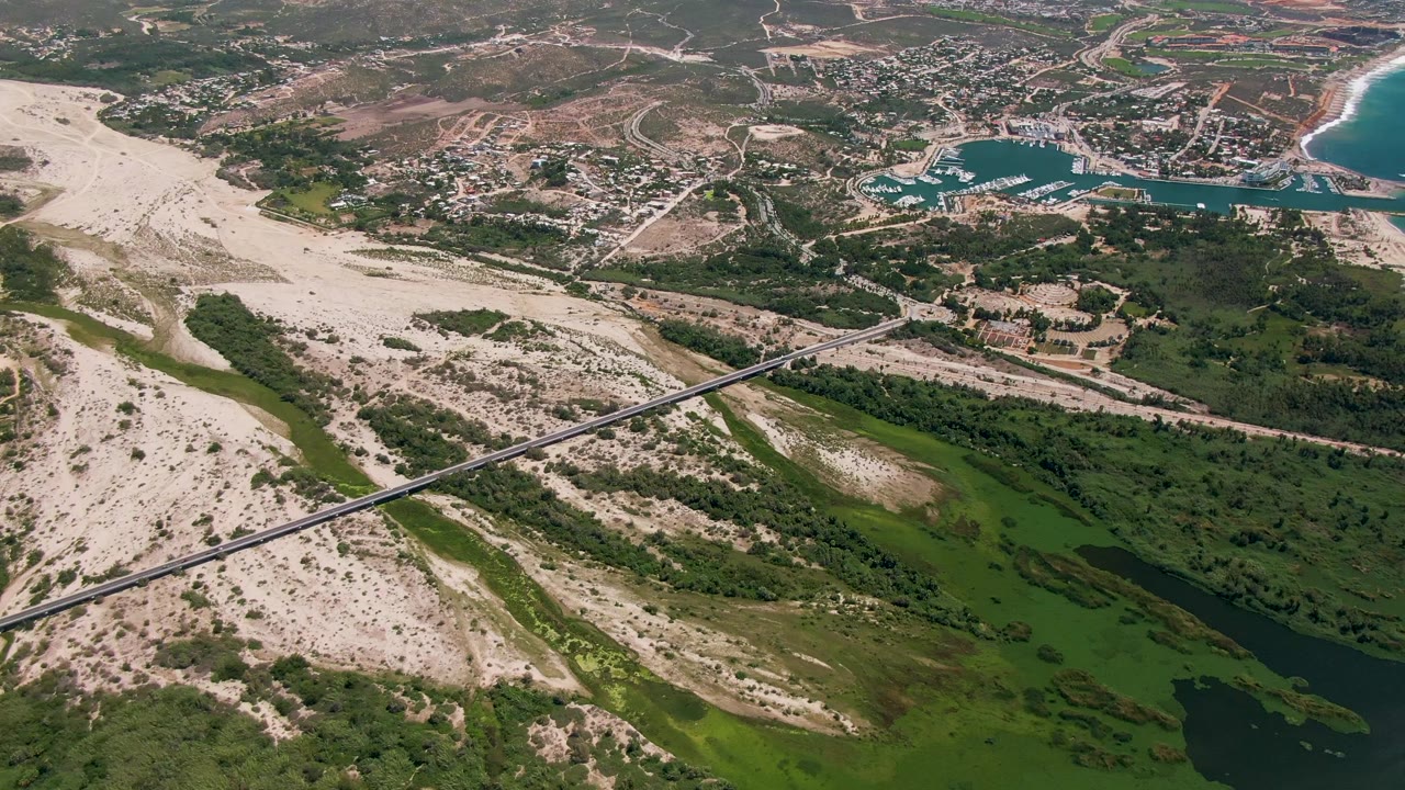 Aerial frame of a sandy river wash, green channel, diagonal bridge, town, and coastline.