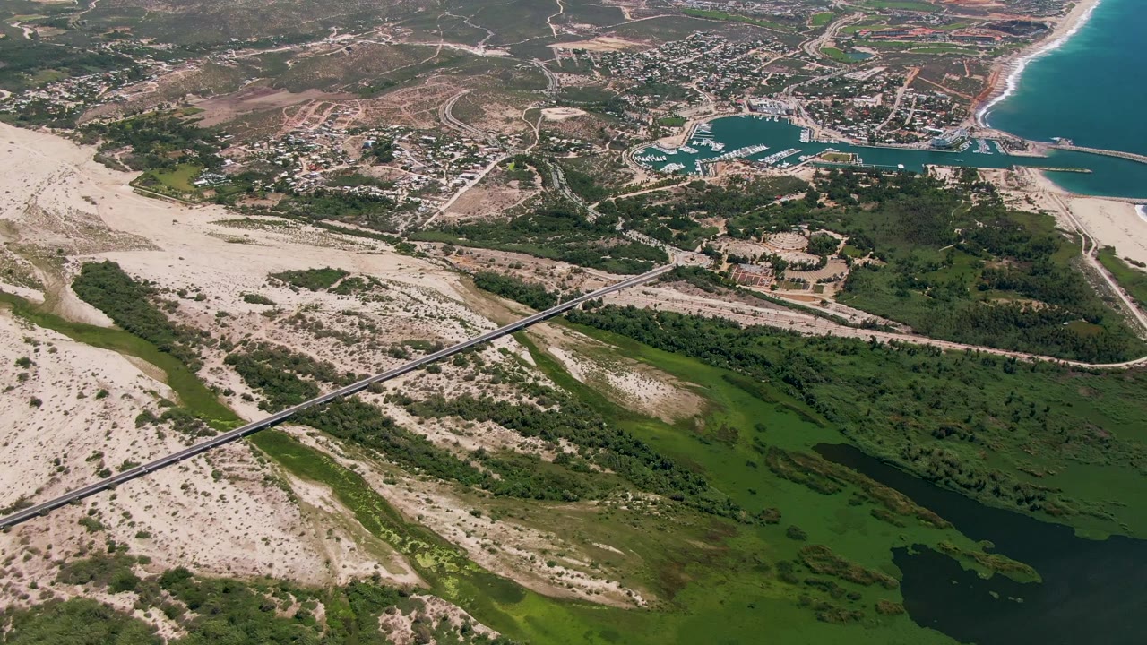 Opening aerial frame over San Jose del Cabo showing a bridge across green estuary water, pale sand dunes, marina basins, beach, ocean, and no visible people.