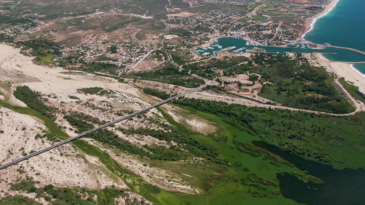 Aerial frame in San Jose del Cabo with the bridge running diagonally over the estuary, dune fields below, marina and town above, and no visible people.