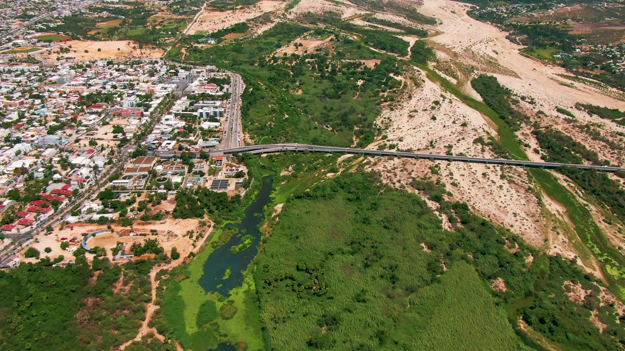 Aerial frame over San Jose Del Cabo showing a highway bridge, green drainage vegetation, residential blocks, sandy hills, and no visible people.