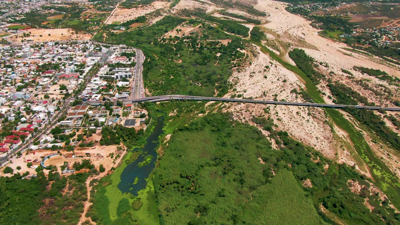 San Jose Del Cabo aerial frame showing light traffic on the bridge, white-roofed buildings, green hills, sandy slopes, and no visible people.