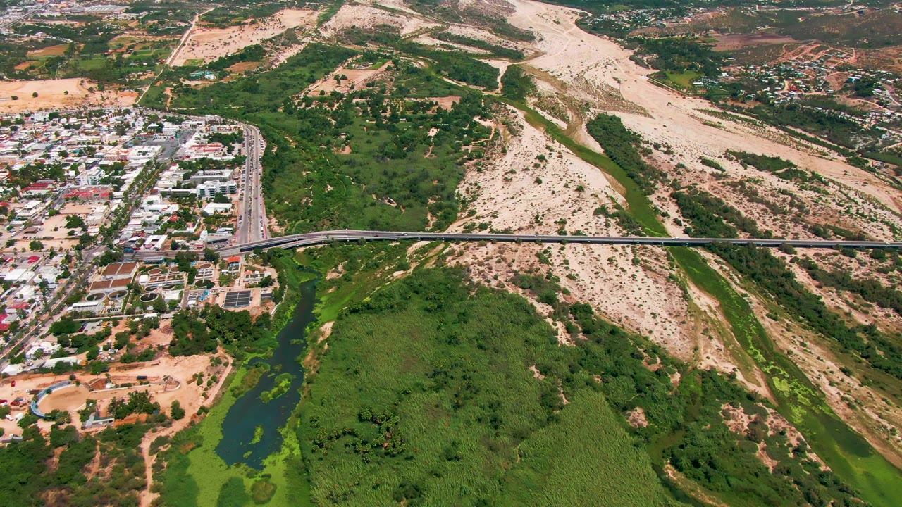 Aerial frame from San Jose del Cabo showing a long bridge over green river vegetation, urban blocks at left, dry hills at right, and no visible people.