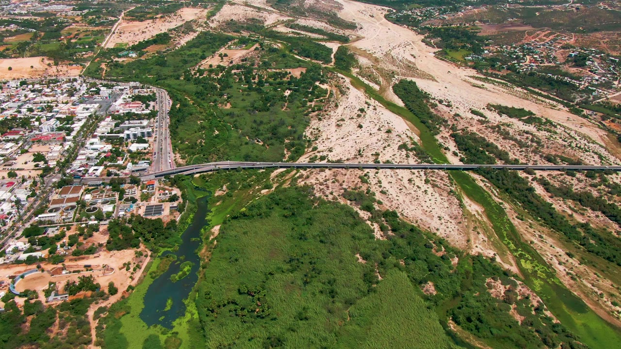 San Jose del Cabo aerial view with the bridge centered across the frame, a developed road grid on the left, green river vegetation below, dry hillside beyond, and no visible people.