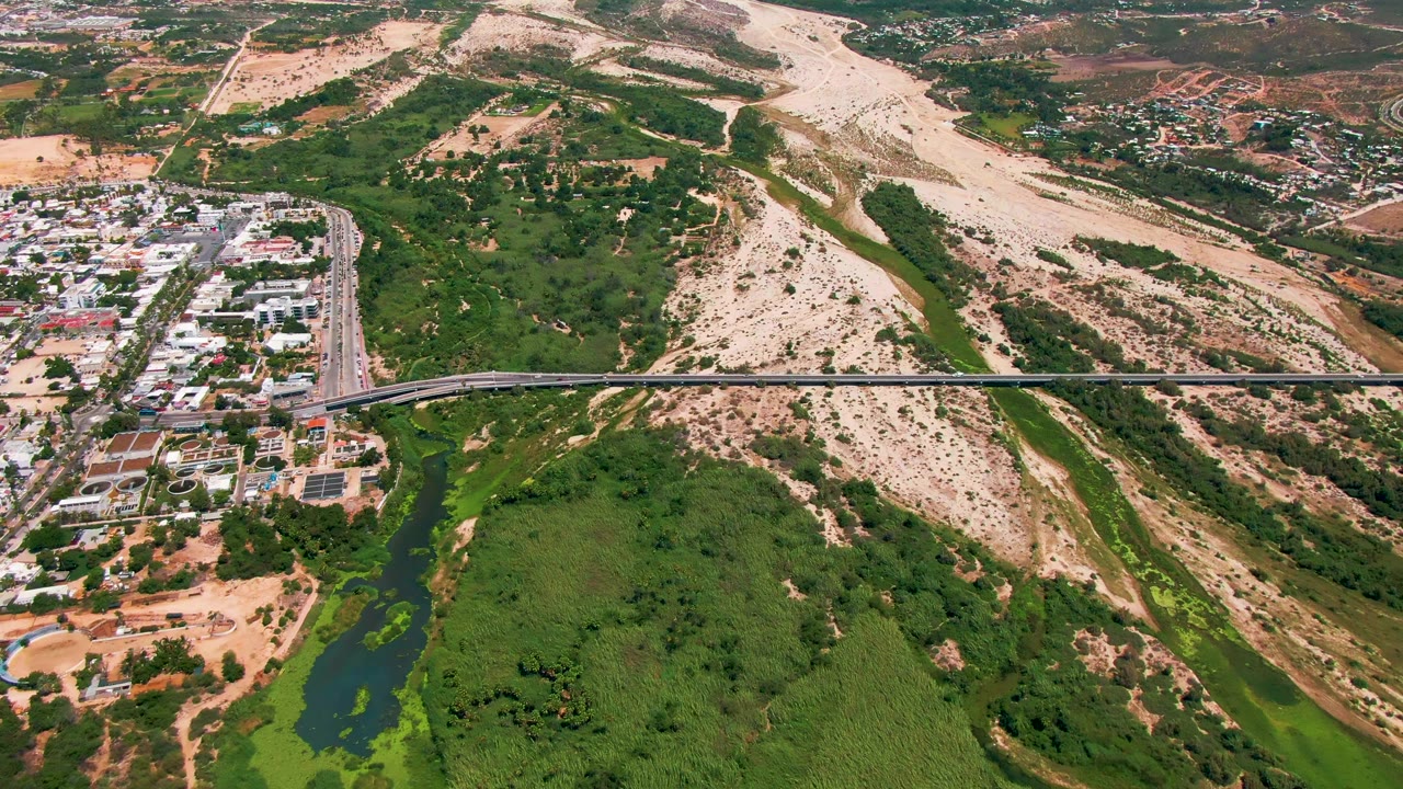 Aerial frame in San Jose del Cabo with the bridge stretching across the lower half, clustered buildings near the road, green water and vegetation below, pale hills above, and no visible people.