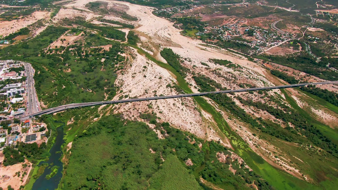 Aerial frame from San Jose del Cabo showing a bridge crossing a river corridor, with buildings and road at the left edge, sandy hills beyond, and no visible people.