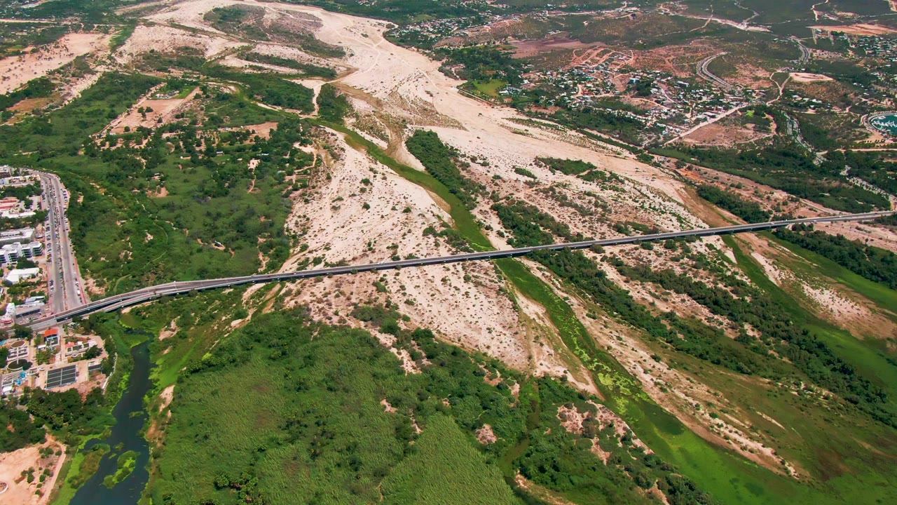 Aerial view in San Jose del Cabo with the bridge centered across green river growth, pale sandy slopes in the background, a road and buildings on the left, and no visible people.