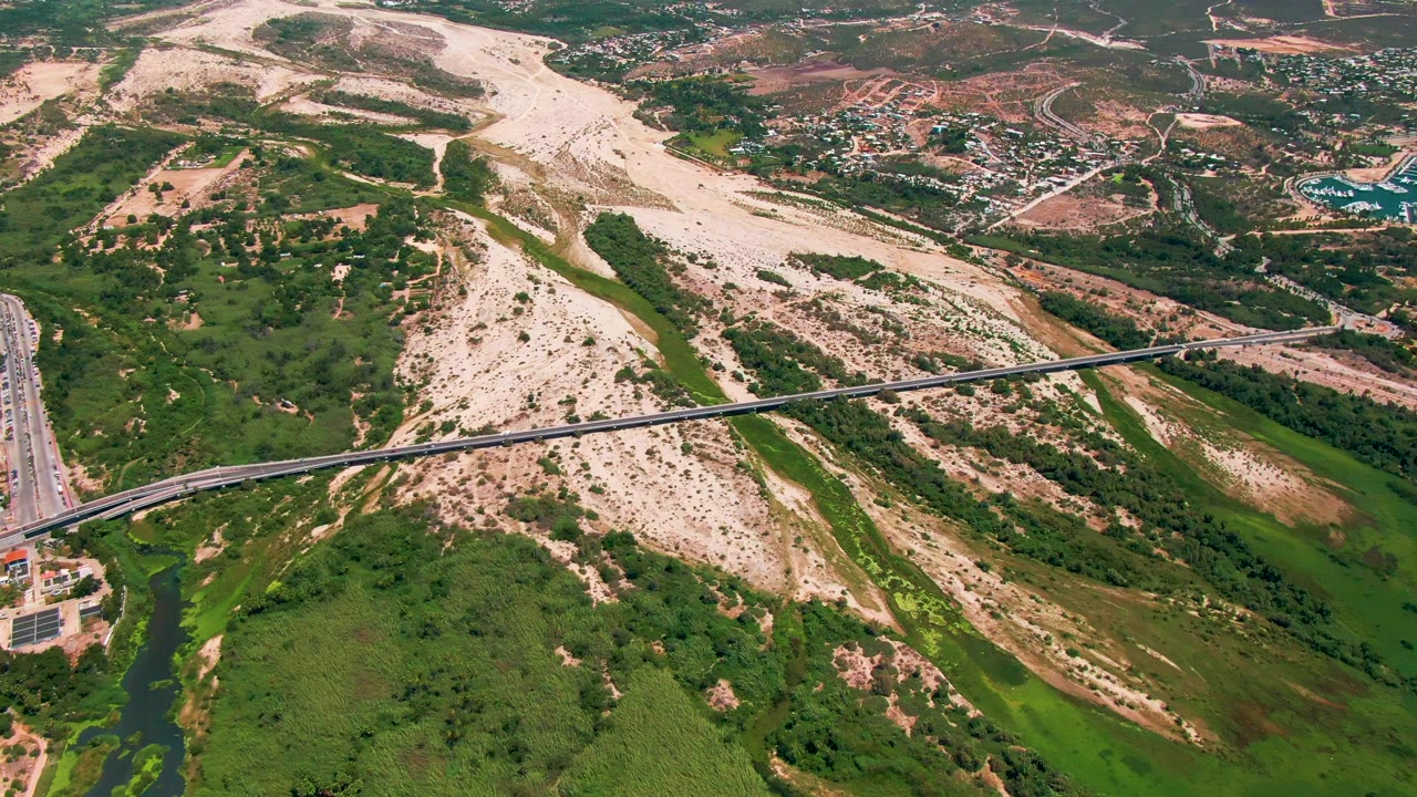 Aerial frame from San Jose del Cabo with more of the green river corridor visible below the bridge, dry sandy ridges behind it, roadside buildings at the far left, and no visible people.