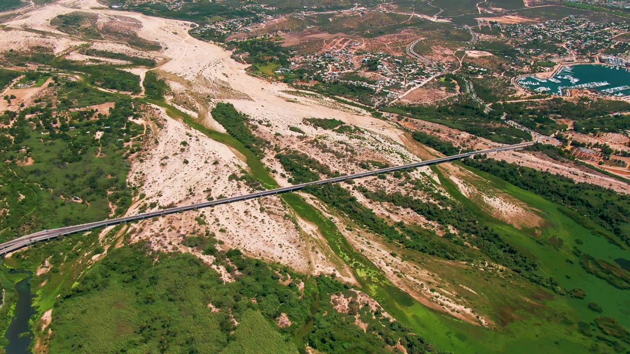 High aerial frame in San Jose del Cabo, Baja California Sur, with the diagonal bridge crossing the valley and a broad dry riverbed stretching toward distant buildings, with no visible people.