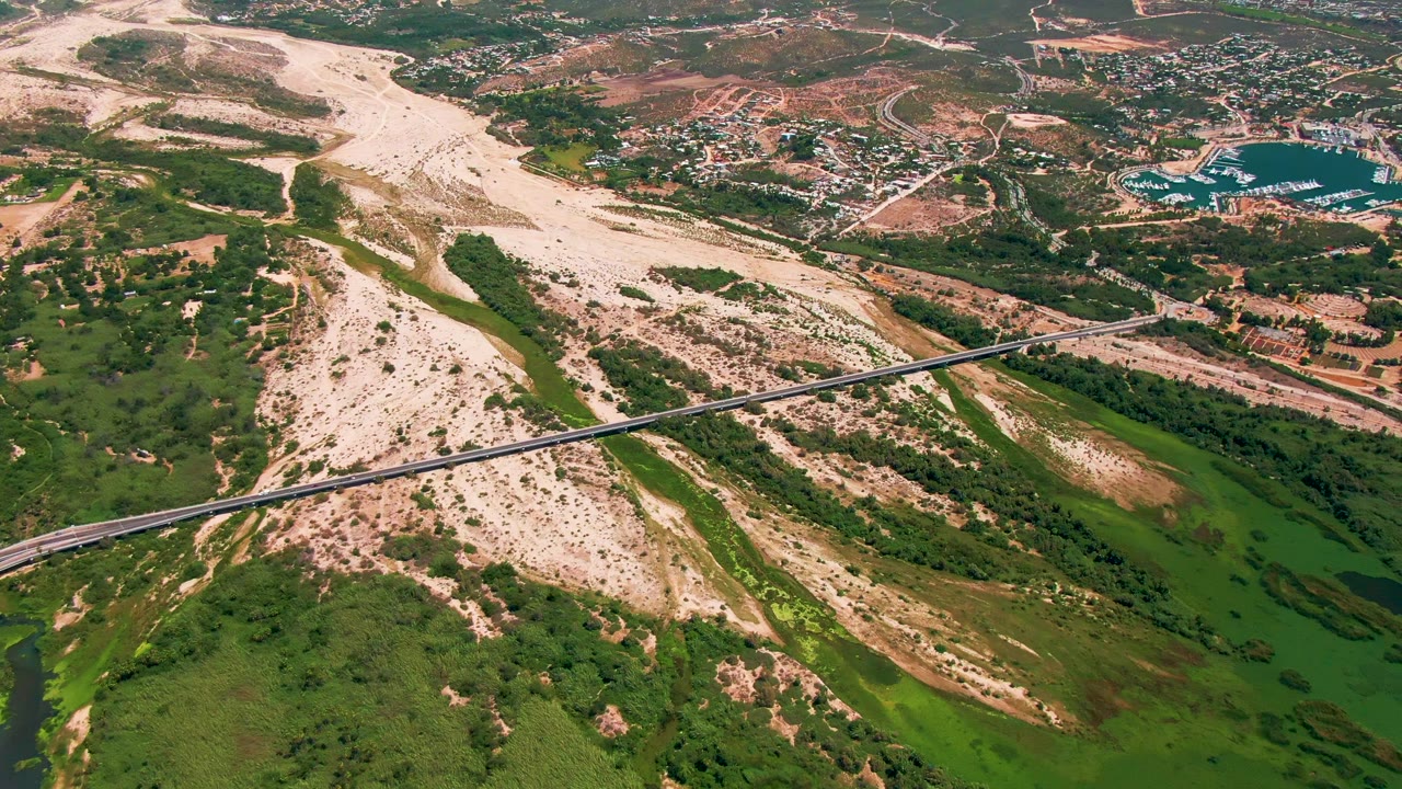 Drone-style aerial view in San Jose del Cabo, Baja California Sur, with the bridge spanning green lowland vegetation and pale terrain, and a turquoise water area near buildings in the background, with no visible people.