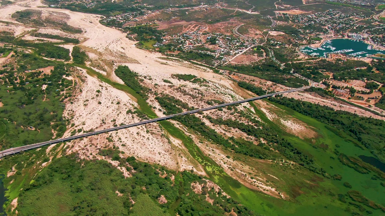 Aerial frame in San Jose del Cabo, Baja California Sur, showing the bridge over green water and vegetation, a pale riverbed across the valley, and dry hills with buildings beyond, with no visible people.