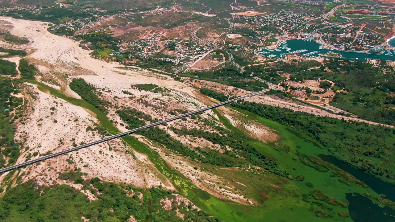 Second aerial frame over San Jose Del Cabo, Mexico, with the bridge spanning the sandy wash, green marshy patches and dark water below, coastal water and town roads in the distance, and no visible people.