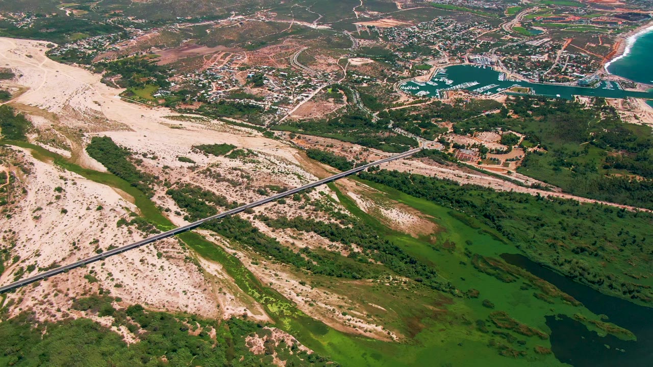 Fourth frame over San Jose Del Cabo, Mexico, showing the bridge crossing a broad sandy channel, dense green vegetation and dark water in the lower right, town buildings beyond, and no visible people.