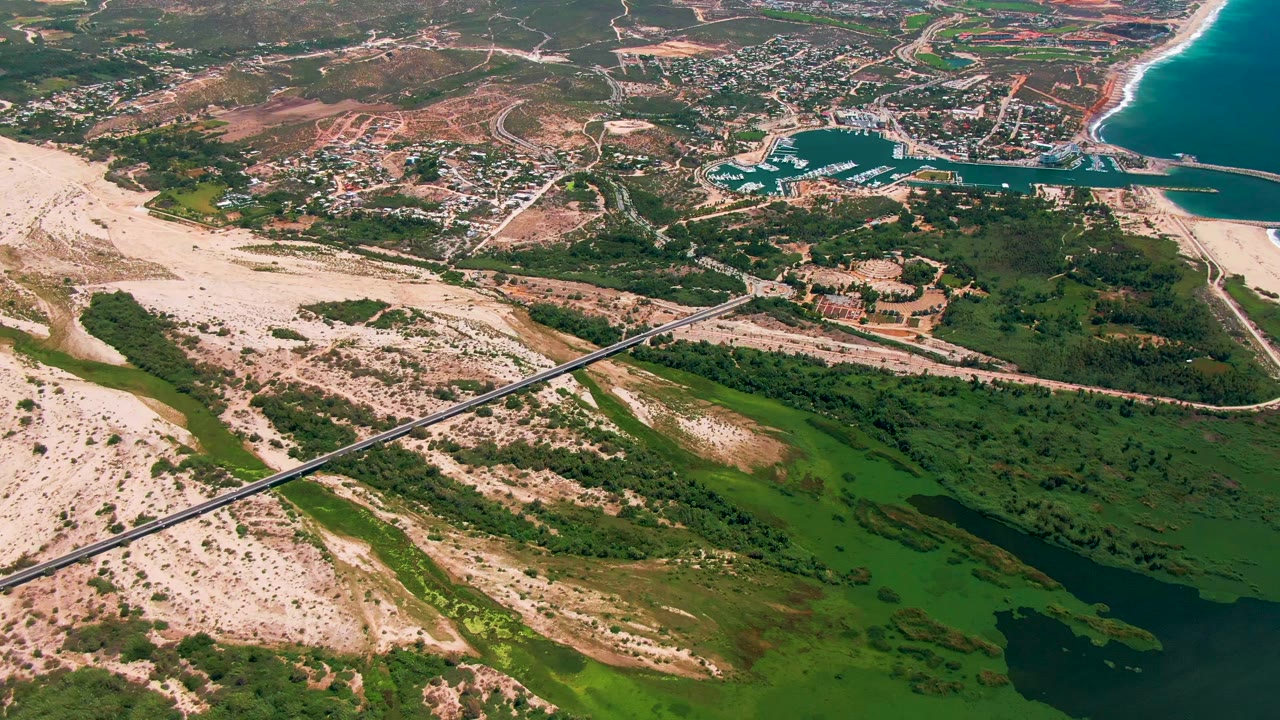 Opening aerial frame over San Jose Del Cabo, Mexico, with a diagonal bridge crossing pale sand and green wetlands toward a marina, beach, town, and blue ocean, with no visible people.