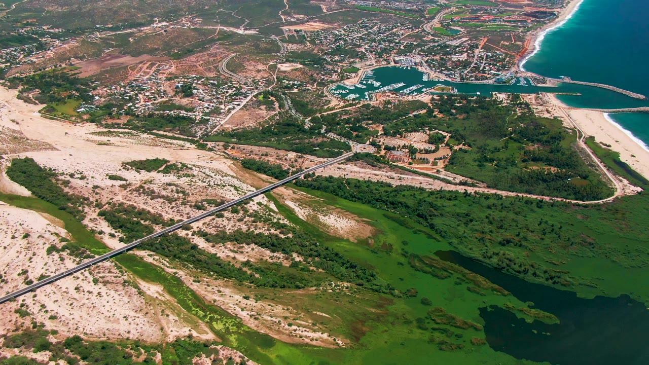 San Jose Del Cabo aerial view with the marina and breakwaters near the upper center, the beach and ocean at right, and the bridge crossing sandy and green terrain below, with no visible people.