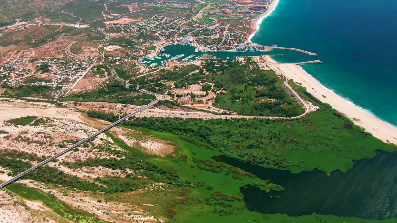 Aerial frame of San Jose Del Cabo with turquoise marina water, boats inside the harbor, pale sand beach, darker ocean water, roadways, green wetland areas, and no visible people.