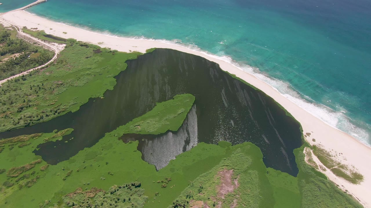 Aerial view in San Jose del Cabo with gentle surf on the diagonal beach, dark lagoon water in the middle ground, and a green cliff mass lower in frame with no visible people.