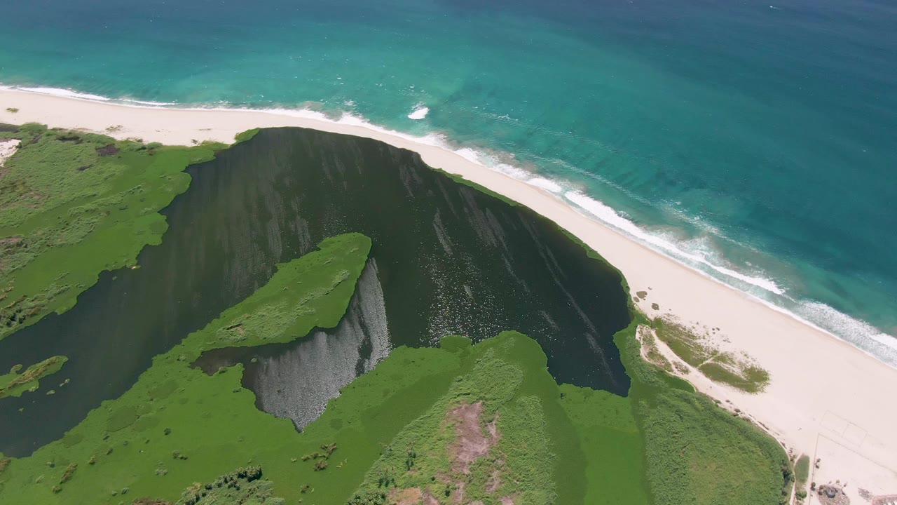 Aerial view of green coastal cliffs above a white beach and turquoise ocean.